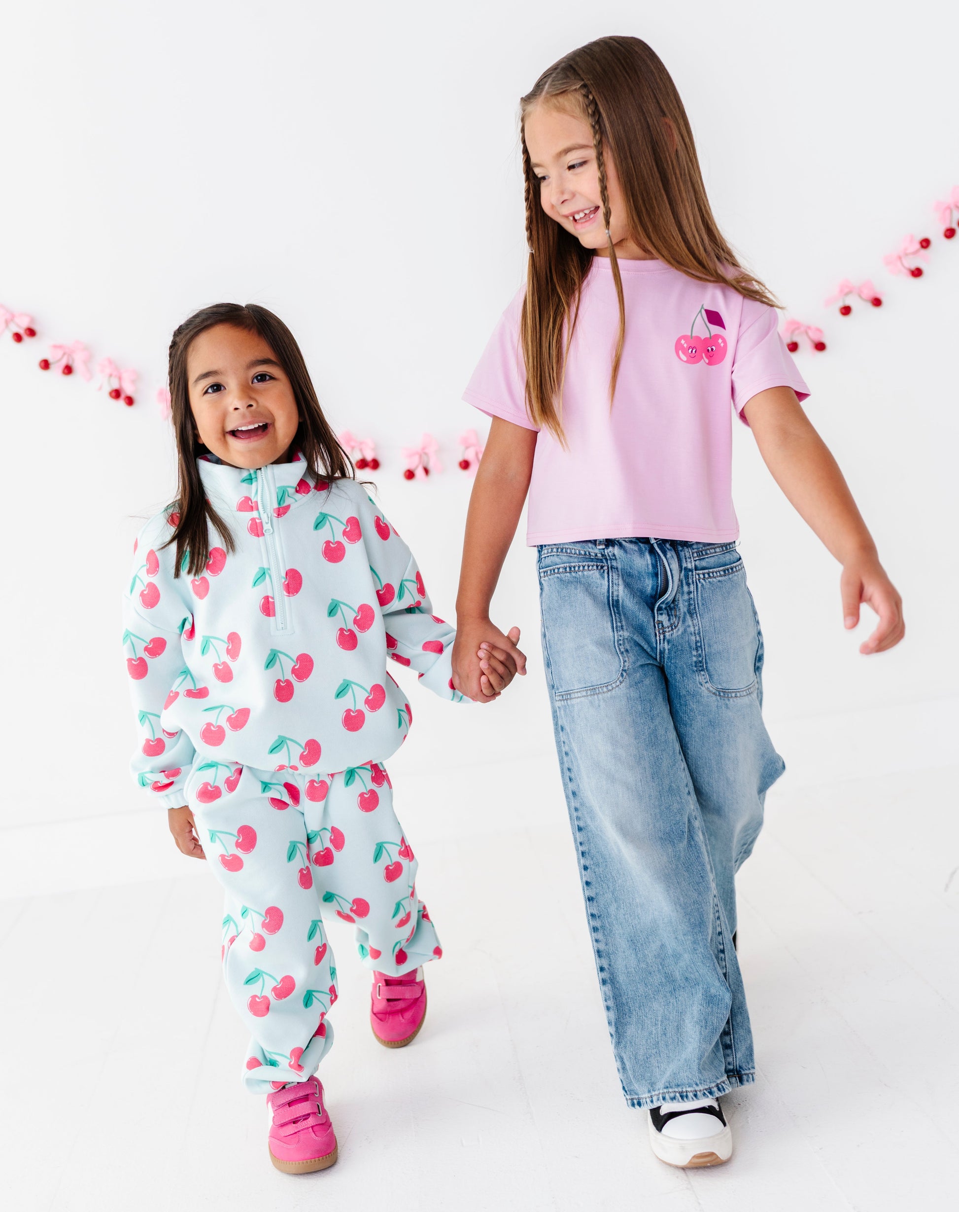 Two young girls walking hand-in-hand in a bright white studio. The girl on the left wears a light blue cherry-patterned tracksuit, and the girl on the right wears a light pink t-shirt with a cherry graphic and wide-leg blue jeans.