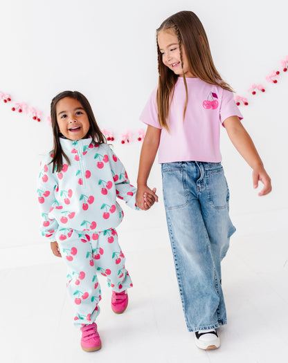 Two young girls walking hand-in-hand in a bright white studio. The girl on the left wears a light blue cherry-patterned tracksuit, and the girl on the right wears a light pink t-shirt with a cherry graphic and wide-leg blue jeans.