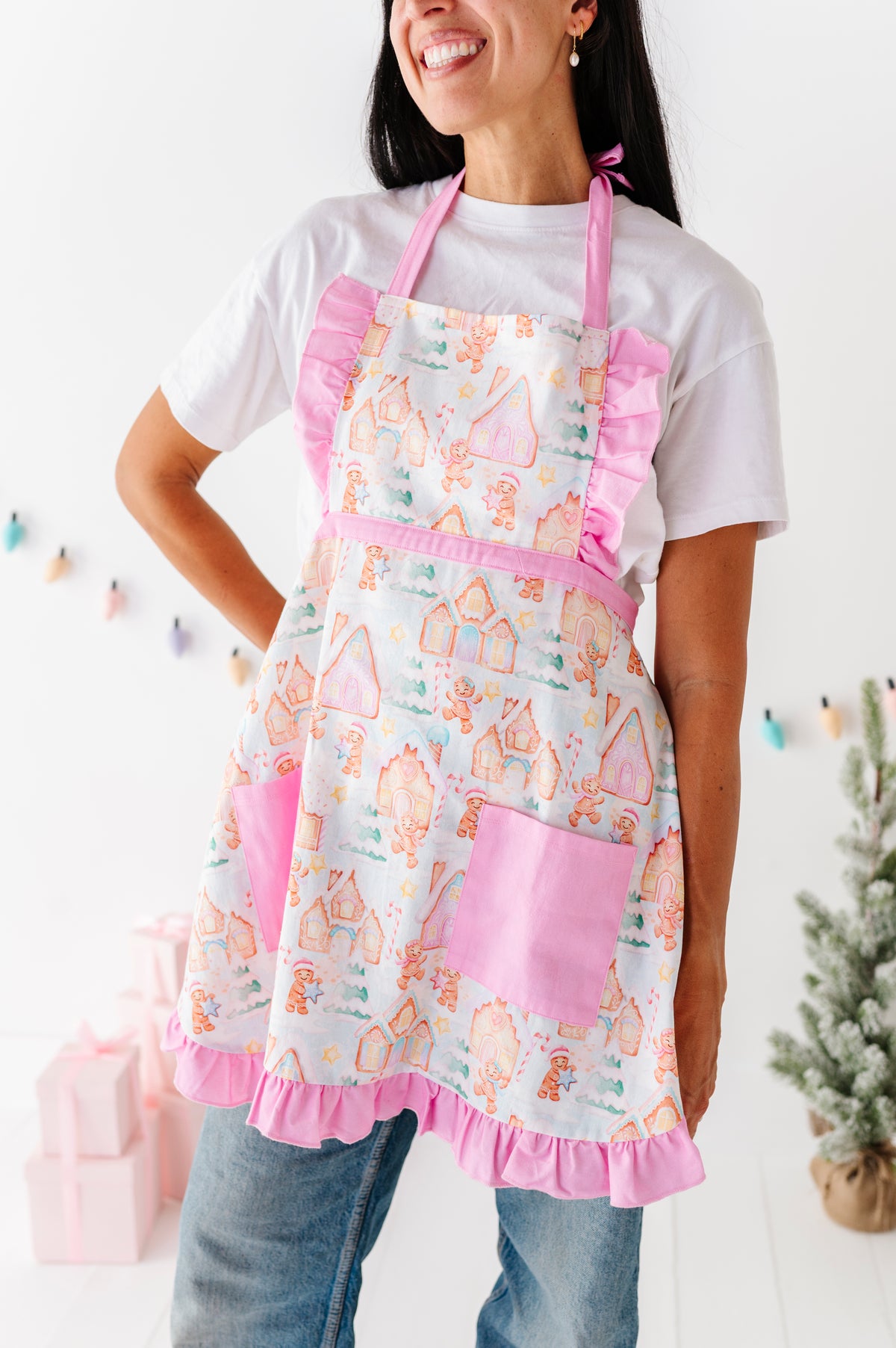 A woman wearing a white apron with pink ruffle trim and a gingerbread house pattern, paired with a white t-shirt and blue jeans, standing in a brightly lit room.