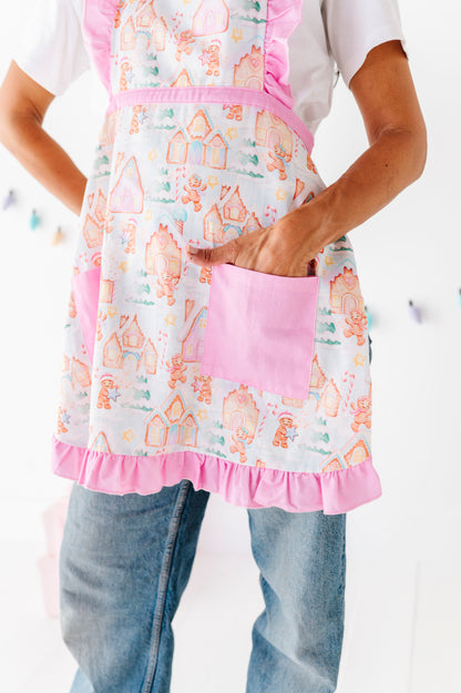 A person wearing a white apron with a pink ruffled trim and a gingerbread house pattern, paired with a white t-shirt and blue jeans.