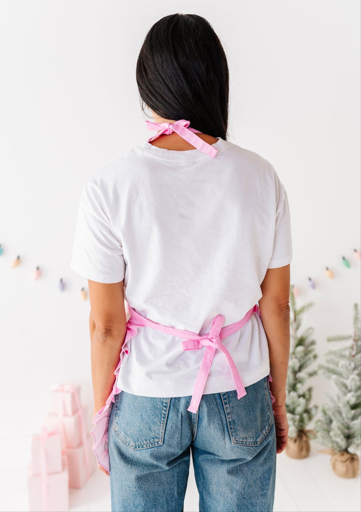 A woman viewed from behind wearing a white T-shirt and blue jeans, with a pink apron tied around her waist and neck, in a bright indoor setting with small decorative trees and gift boxes.