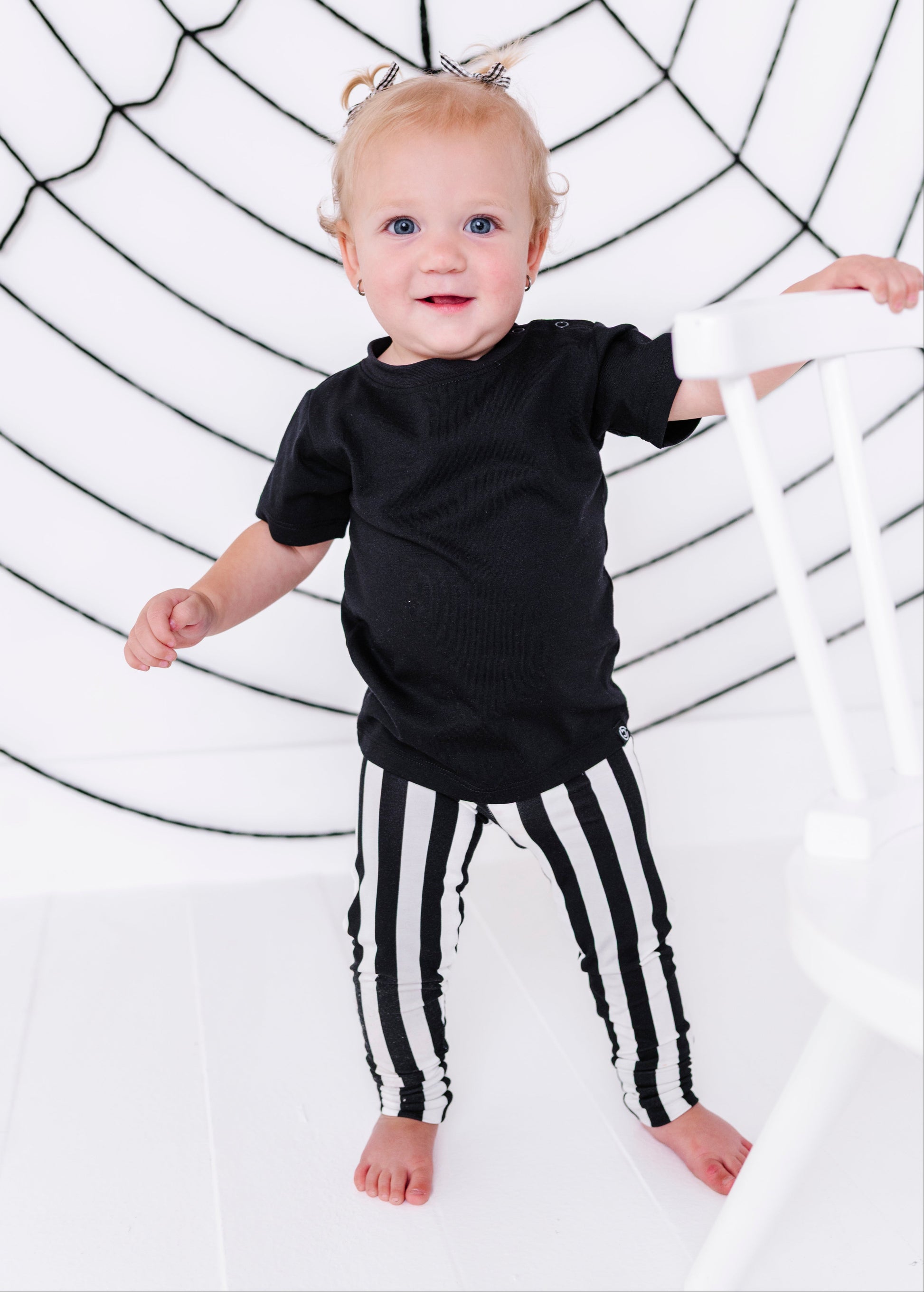 A toddler standing barefoot in a black t-shirt and black-and-white vertically striped pants, holding onto a white chair in front of a white background with a black spiderweb graphic.
