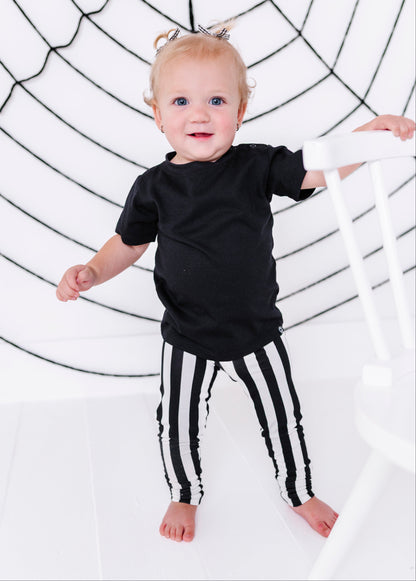 A toddler standing barefoot in a black t-shirt and black-and-white vertically striped pants, holding onto a white chair in front of a white background with a black spiderweb graphic.