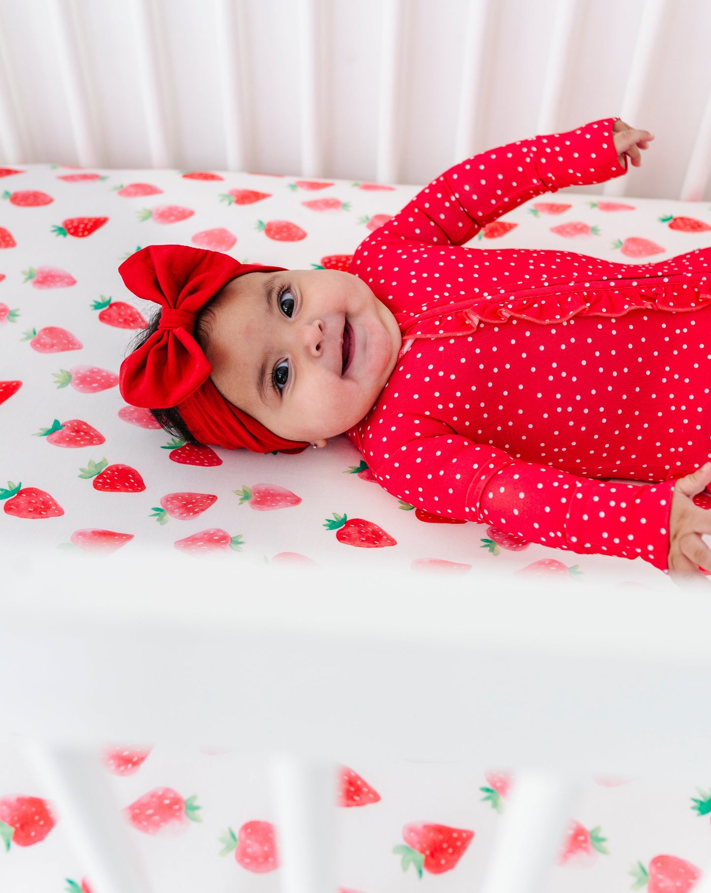 Baby in a red romper with a large bow lying on a strawberry-patterned blanket.