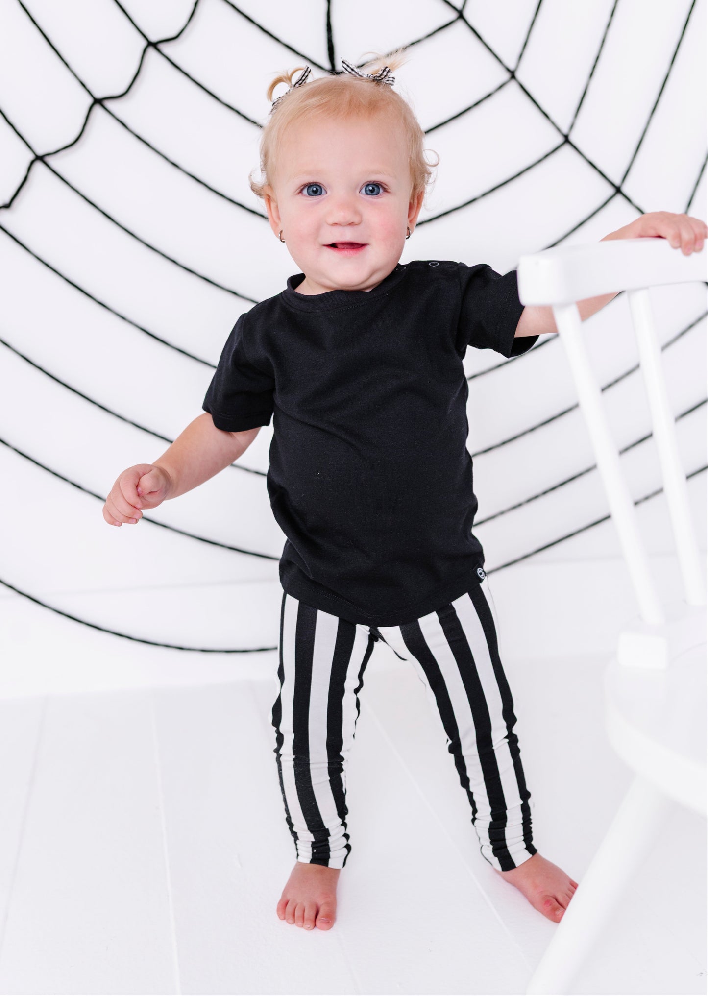 A toddler standing barefoot in a black t-shirt and black-and-white vertically striped pants, holding onto a white chair in front of a white background with a black spiderweb graphic.