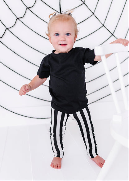 A toddler standing barefoot in a black t-shirt and black-and-white vertically striped pants, holding onto a white chair in front of a white background with a black spiderweb graphic.