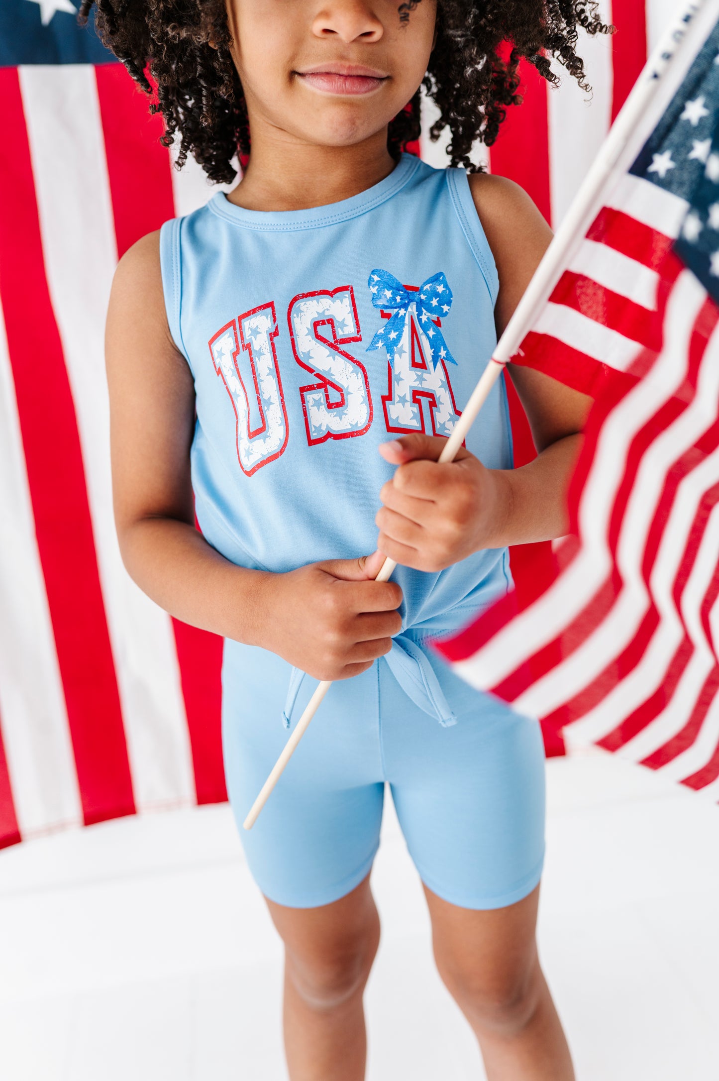 Child wearing a blue bamboo 'USA' tank top and bamboo biker shorts holding an American flag