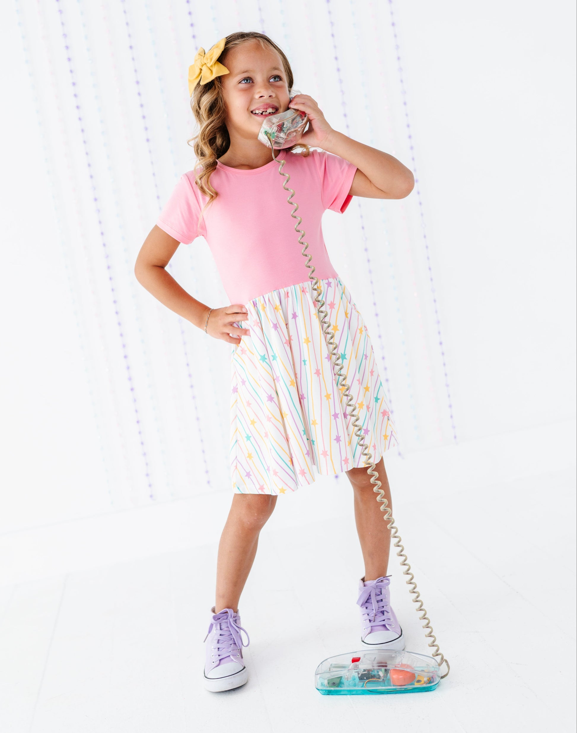 A young girl with a yellow bow in her hair is wearing a pink short-sleeve t-shirt and a white pleated skirt with pastel rainbow stripes. She is holding a retro-style silver rotary telephone receiver and posing with a matching clear plastic rotary phone on the floor against a white background with vertical light patterns.