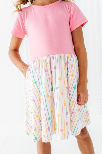 A child wearing a short-sleeve dress with a solid pink bodice and a white skirt patterned with colorful stars and vertical lines.