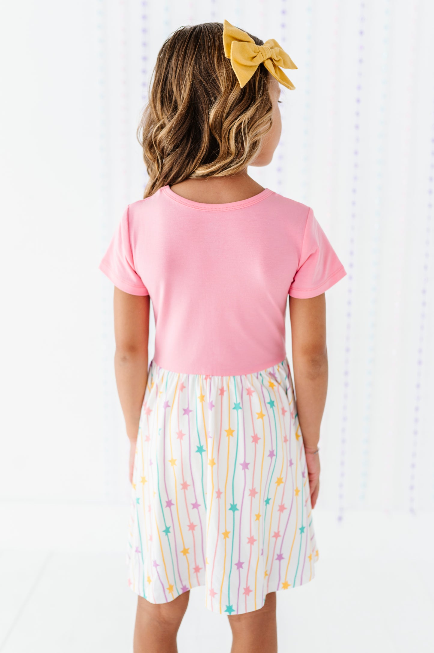 A young girl viewed from behind wearing a short-sleeve pink top and a white skirt with vertical pastel rainbow stripes and colorful stars. She has a yellow bow in her hair.