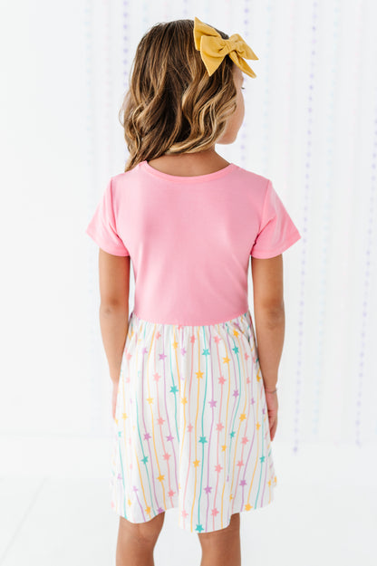 A young girl viewed from behind wearing a short-sleeve pink top and a white skirt with vertical pastel rainbow stripes and colorful stars. She has a yellow bow in her hair.
