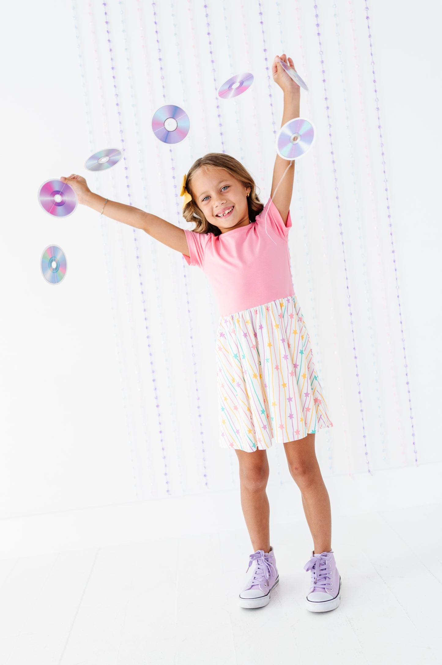 A young girl with blonde hair in a ponytail is smiling and posing with floating iridescent CD-like discs. She is wearing a pink t-shirt, a white skirt with pastel rainbow stripes, and light purple sneakers, set against a bright white background with hanging string lights.
