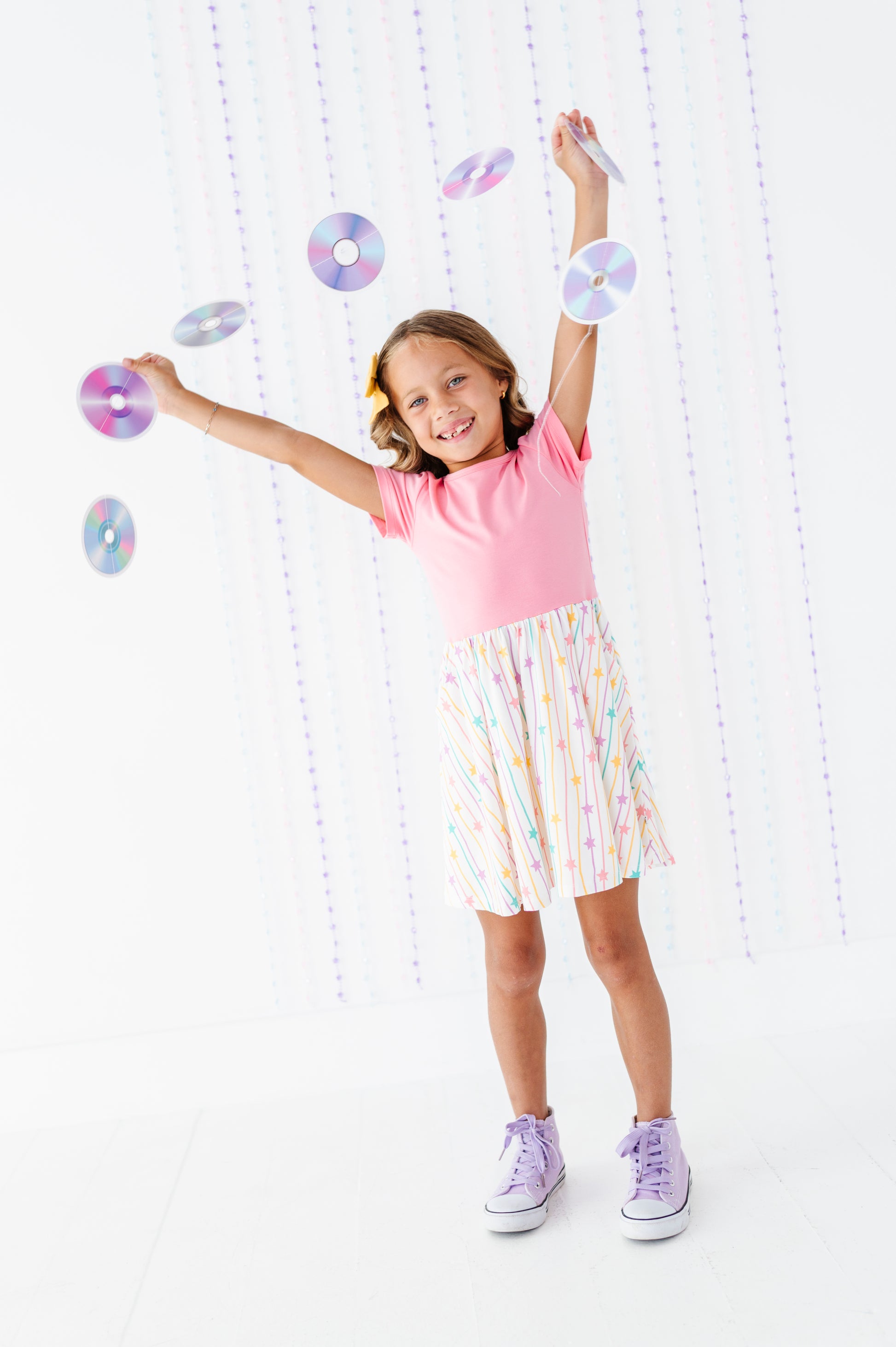A young girl with blonde hair in a ponytail is smiling and posing with floating iridescent CD-like discs. She is wearing a pink t-shirt, a white skirt with pastel rainbow stripes, and light purple sneakers, set against a bright white background with hanging string lights.