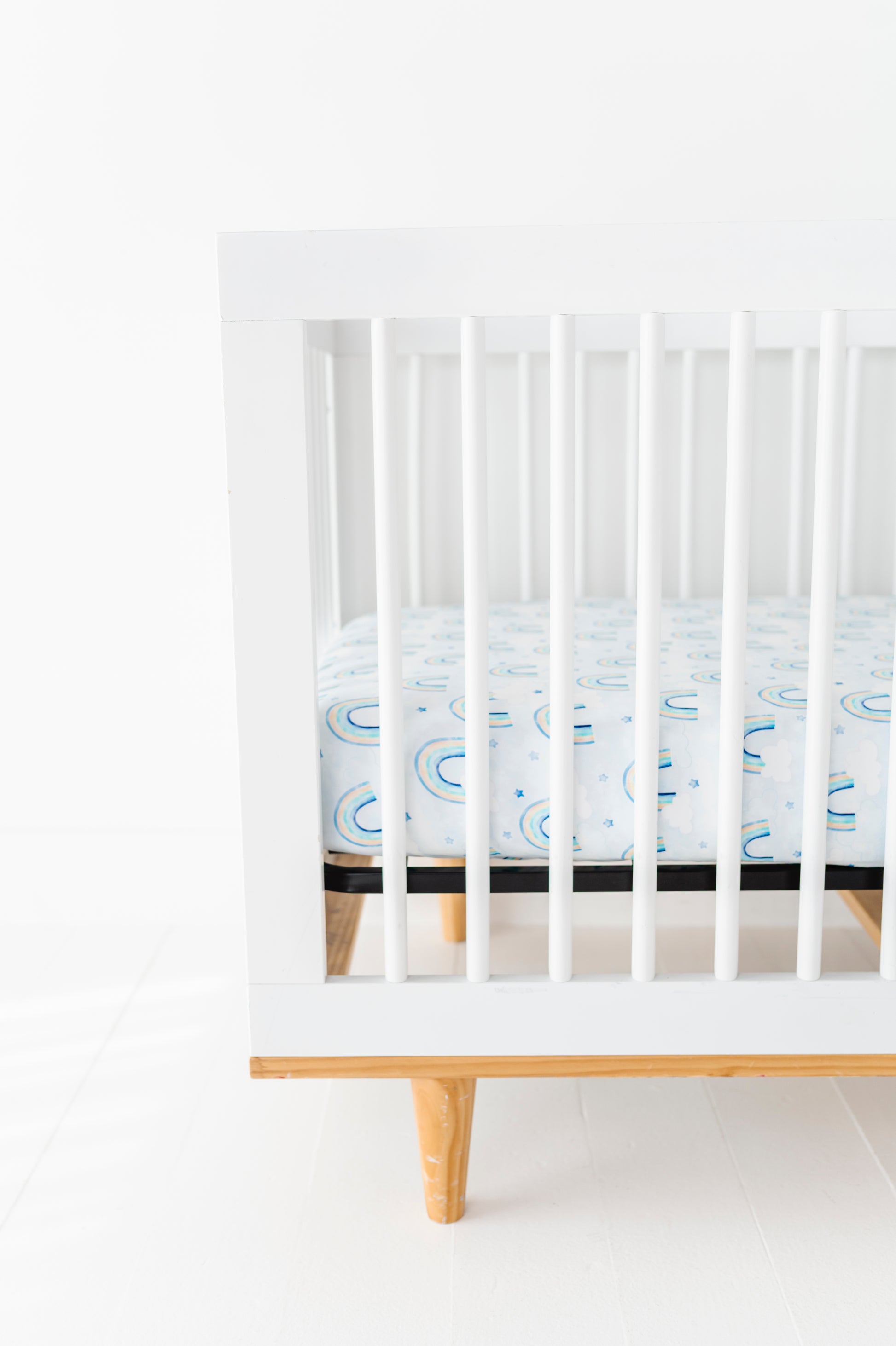 White crib with a mattress featuring a bamboo crib sheet with pastel watercolor rainbows