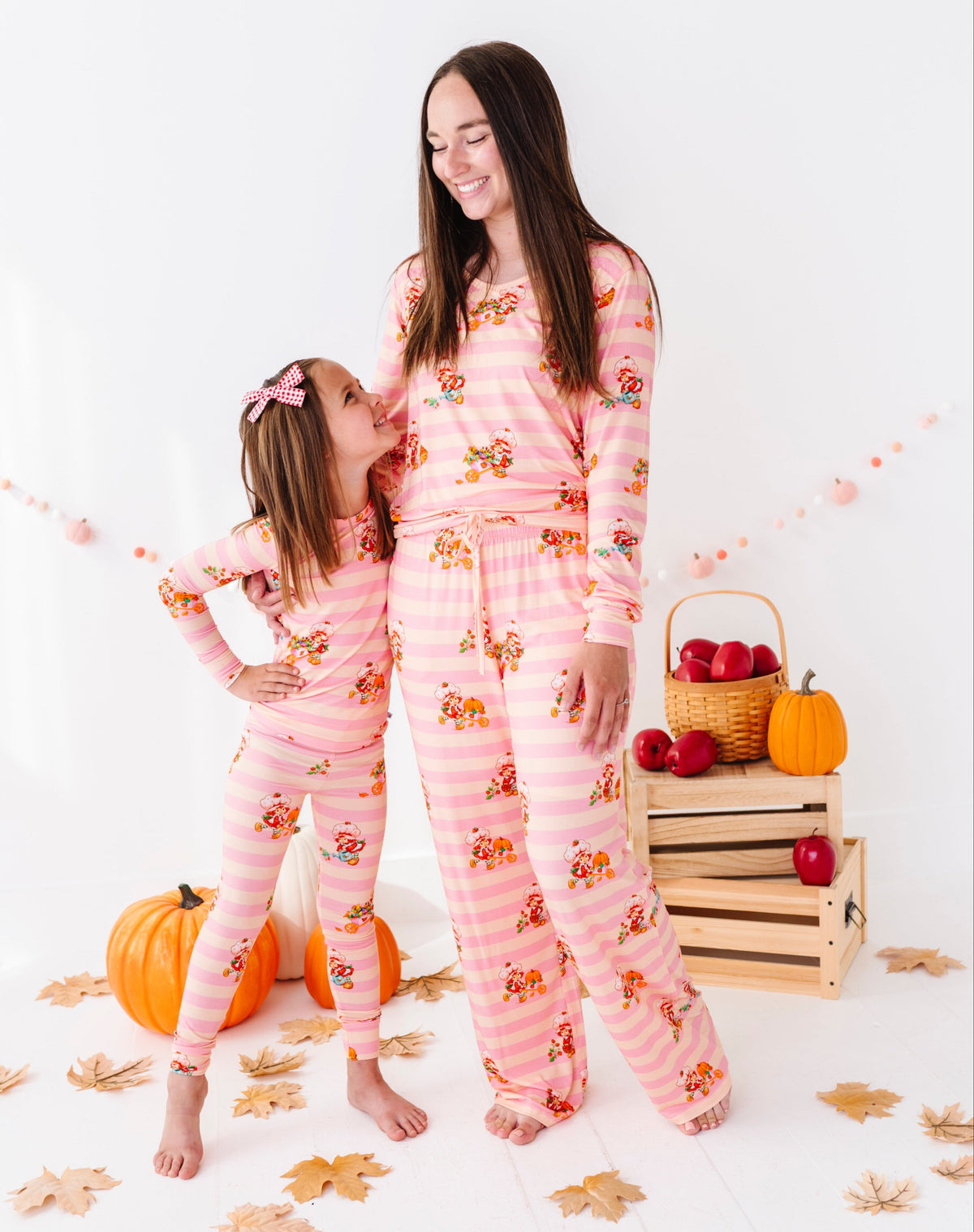 A woman and a young girl wearing matching pink autumn-themed pajamas with a pumpkin and leaf print, standing in a studio decorated with pumpkins, fall leaves, and a wooden crate of red apples.