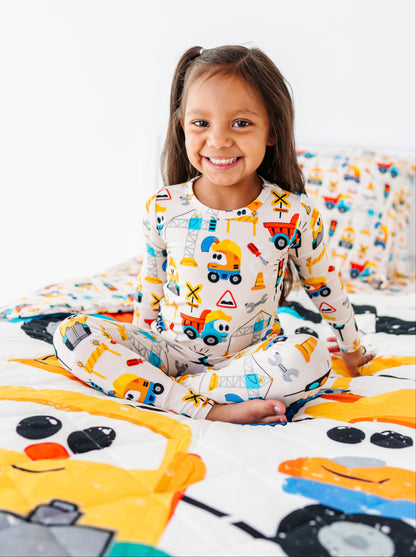 A young girl with dark hair in pigtails sits cross-legged on a bed, smiling at the camera. She is wearing a one-piece pajama set with a colorful construction vehicle print, and the bed features matching printed bedding with cartoon vehicles.