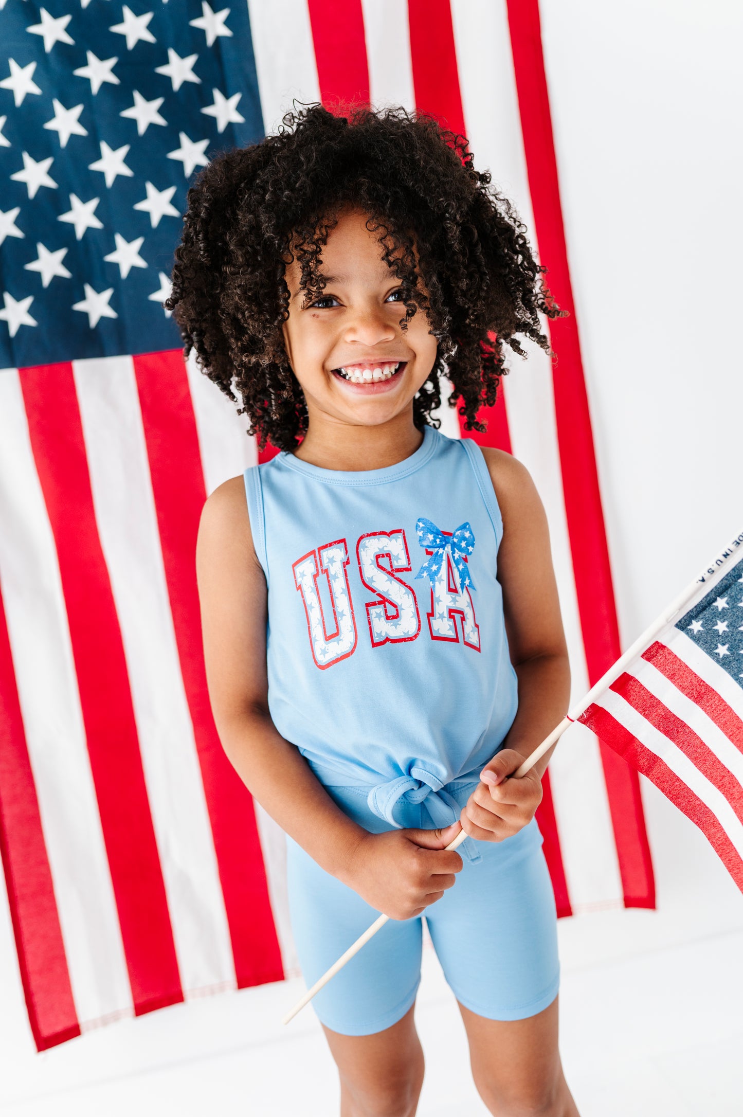 Child wearing a bamboo 4th of July tank top and coordinating bamboo biker shorts, and holding an American flag in front of another American flag.