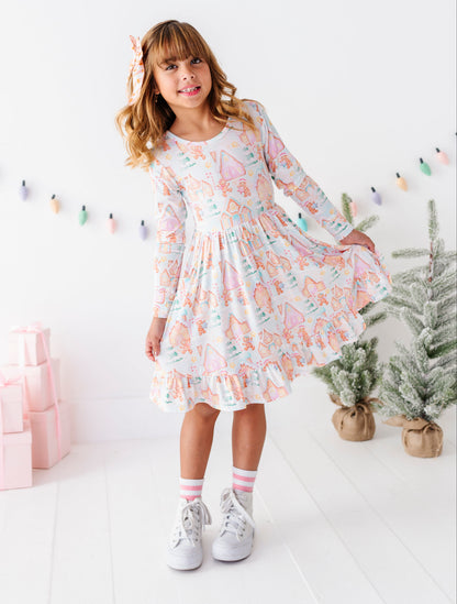 A young girl with blonde hair wearing a long-sleeve white dress with a pastel pink, orange, and blue unicorn pattern, standing in a bright white room decorated with gift boxes, a small tree, and a string of colorful lights.
