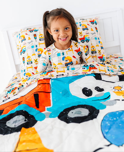 A young girl with dark hair in a ponytail sits in bed smiling, wearing a white long-sleeve pajama shirt with a colorful construction vehicle pattern. The bedding set features a matching patterned pillowcase and a quilted comforter with a large cartoon panda face and construction vehicles.