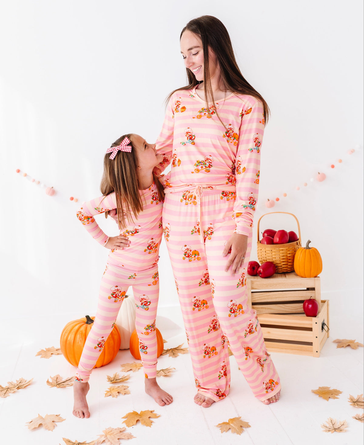 A woman and a young girl wearing matching pink striped pajamas with a fall leaf print, posed in a studio setting with pumpkins, apples, and autumn leaves.