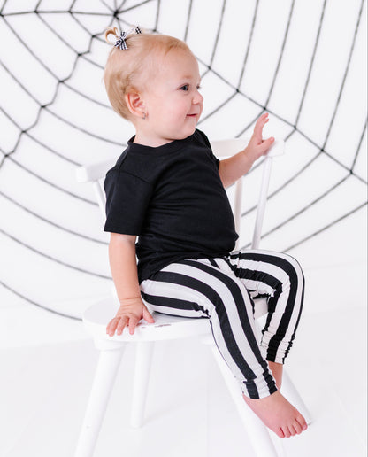 A baby sitting on a white chair, wearing a black t-shirt and black and white striped pants, with a spiderweb backdrop behind them.