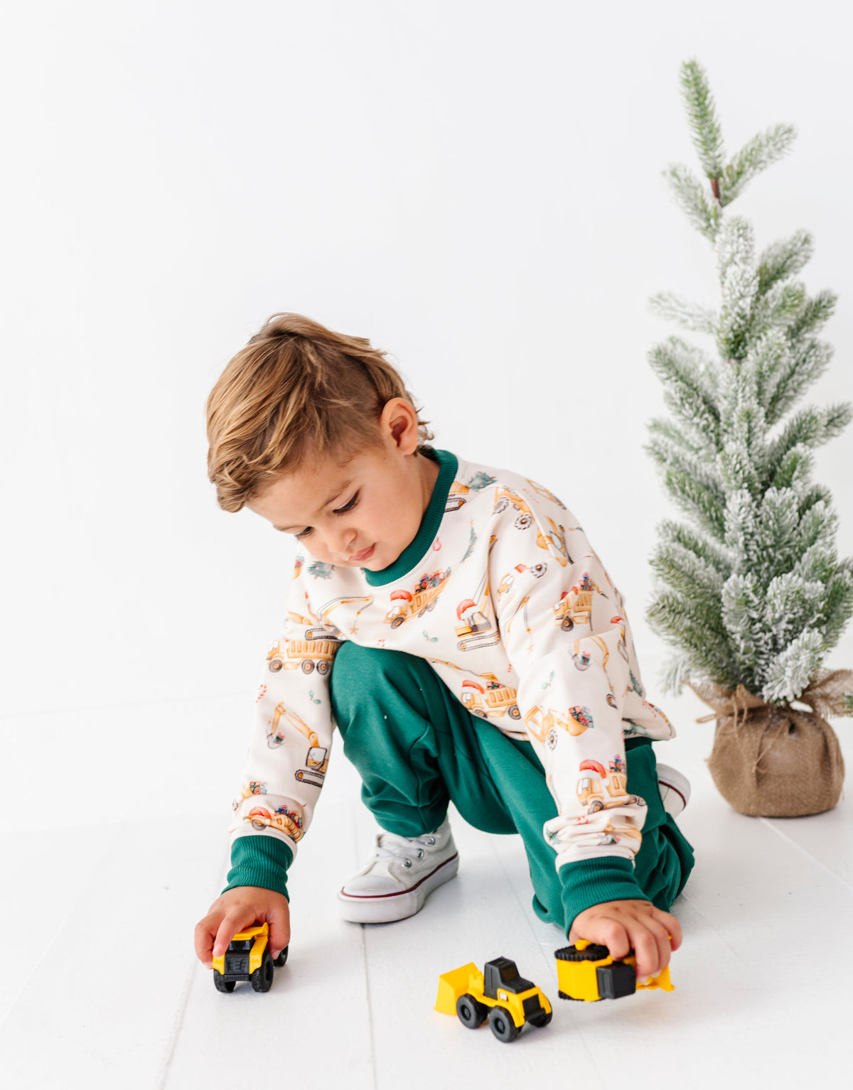 A young boy wearing a construction crew crewneck and matching green joggers, playing with yellow toy trucks on a white floor next to a small decorated tree.
