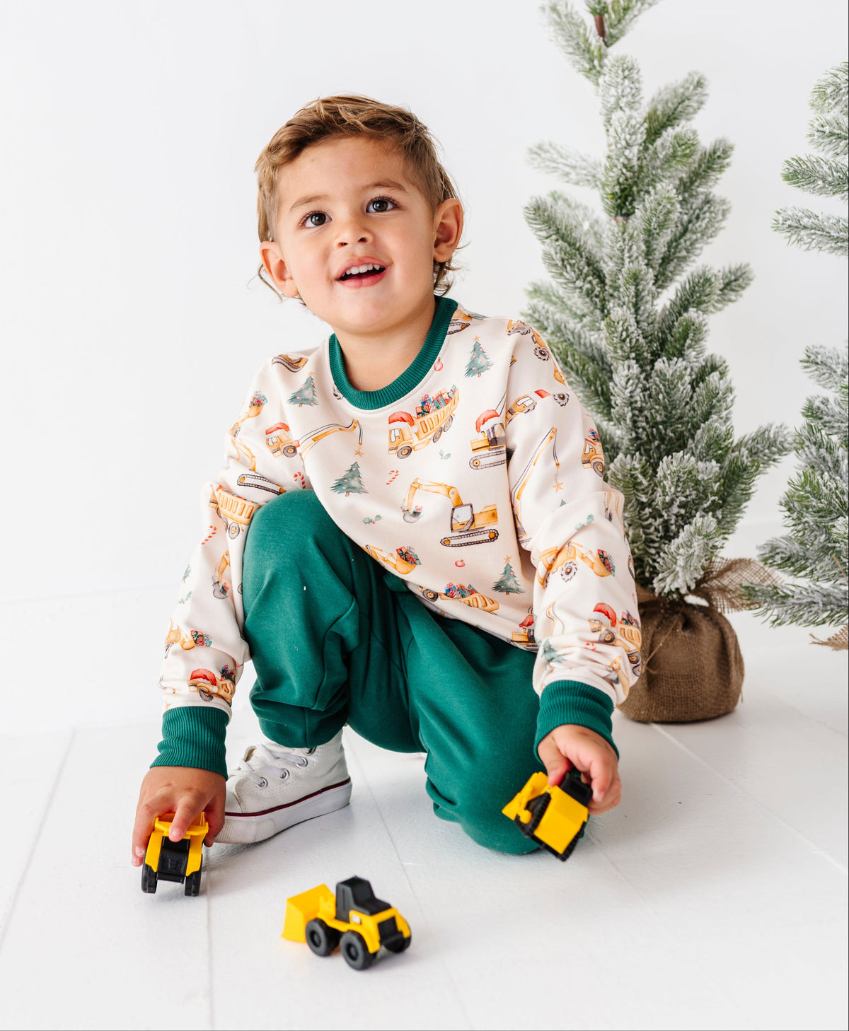 A young boy wearing a construction-themed crewneck and jogger set crouches on a white floor, holding yellow toy trucks. The pajamas feature a cream background with colorful cartoon construction vehicles, with dark green cuffs and collar.