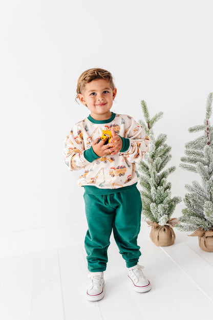 A young boy stands in a bright white studio wearing a holiday-themed sweatshirt and sweatpants set. The sweatshirt is cream with a pattern of Christmas trees, ornaments, and sleighs, paired with solid green cuffs and collar, and matching green sweatpants.