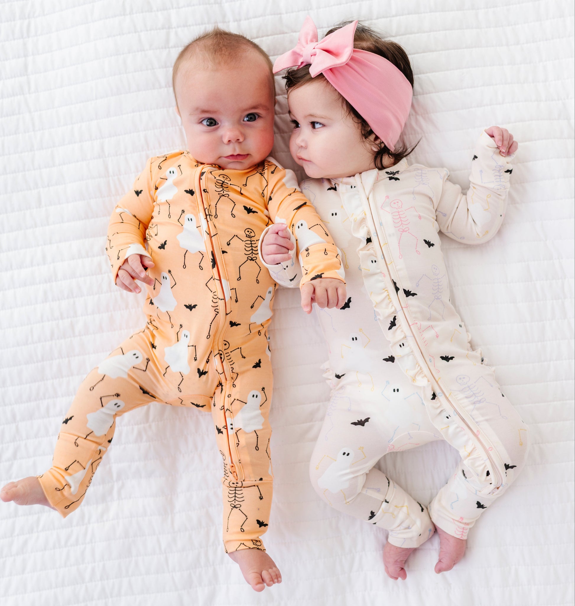 Two babies wearing halloween-themed onesies on a white textured surface