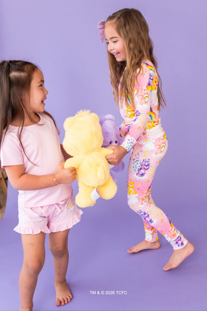 Two children in colorful Care Bears™ outfits holding a plush toy against a purple background