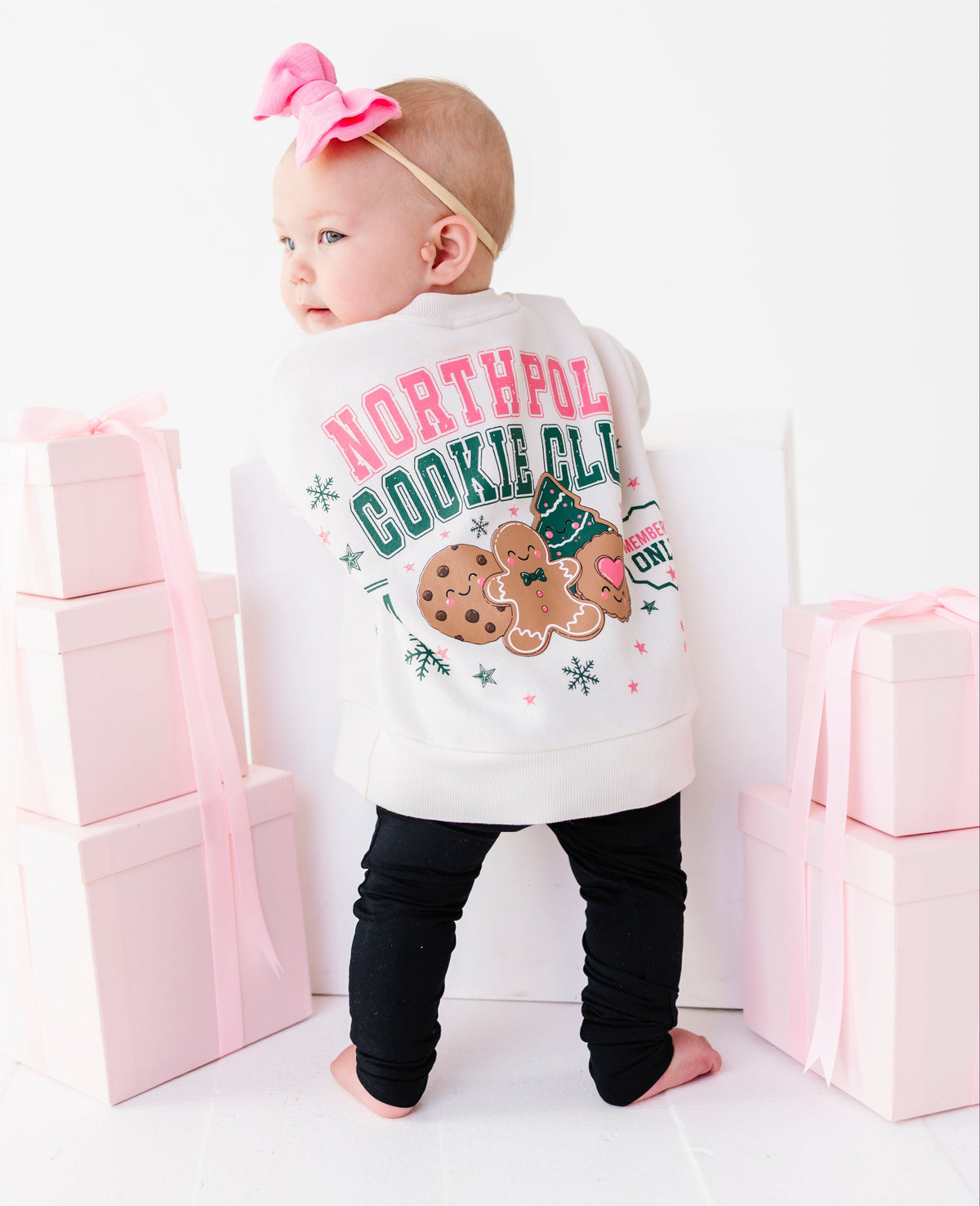 A baby standing with their back to the camera, wearing a crewneck with a colorful 'NORTH POLE COOKIE CLUB' graphic and black leggings. The baby is framed by stacks of pink gift boxes on a clean white background.