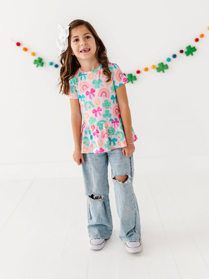 Young girl wearing a colorful bamboo cotton top and ripped jeans standing against a white background with St. Patrick's Day decorative string lights.