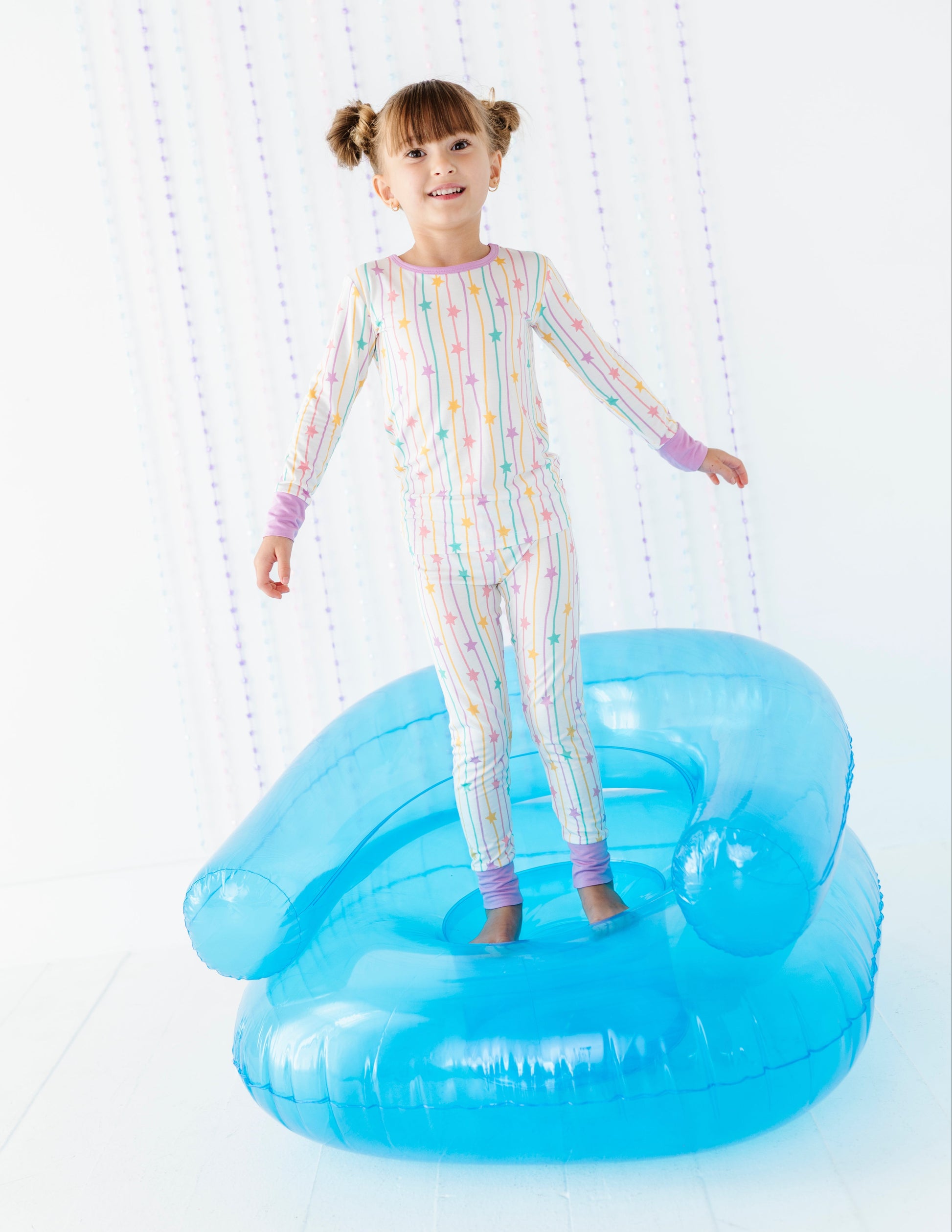 A young girl with pigtails stands on a large blue novelty chair in a bright white room, wearing a pastel rainbow-striped long-sleeve pajama set.