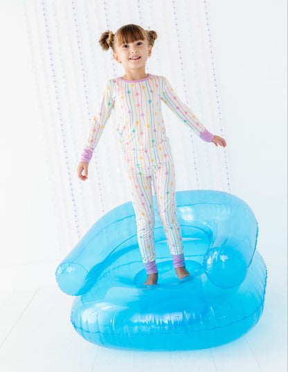 A young girl with pigtails stands on a large blue novelty chair in a bright white room, wearing a pastel rainbow-striped long-sleeve pajama set.