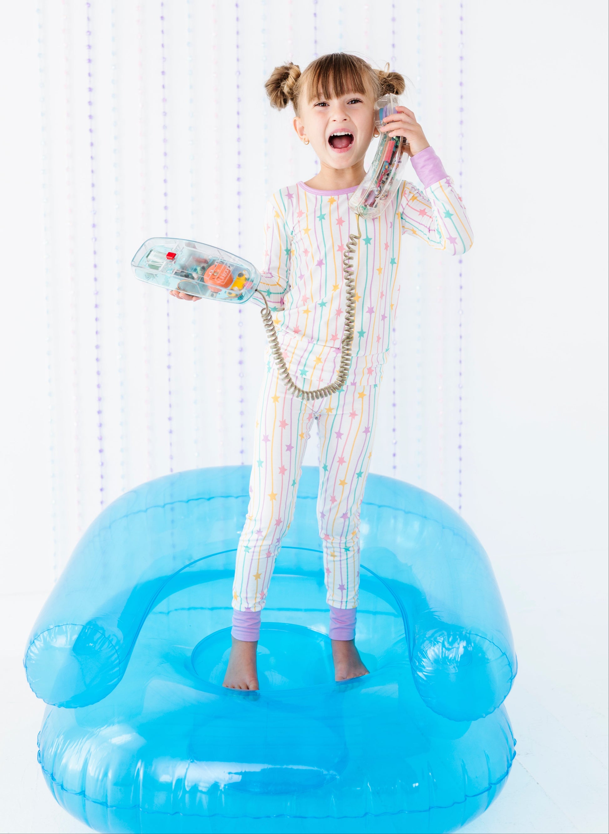 A young girl with pigtails stands barefoot on a translucent blue inflatable chair, wearing a pastel rainbow-striped pajama set and holding a retro-style toy telephone to her ear.