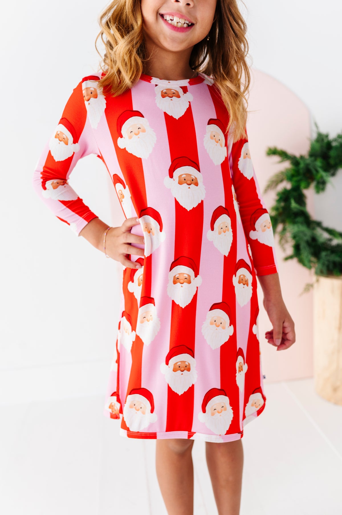 A young girl wearing a long-sleeve bamboo gown with a festive Santa Claus pattern, featuring alternating vertical stripes of red and light pink with Santa faces and beards.
