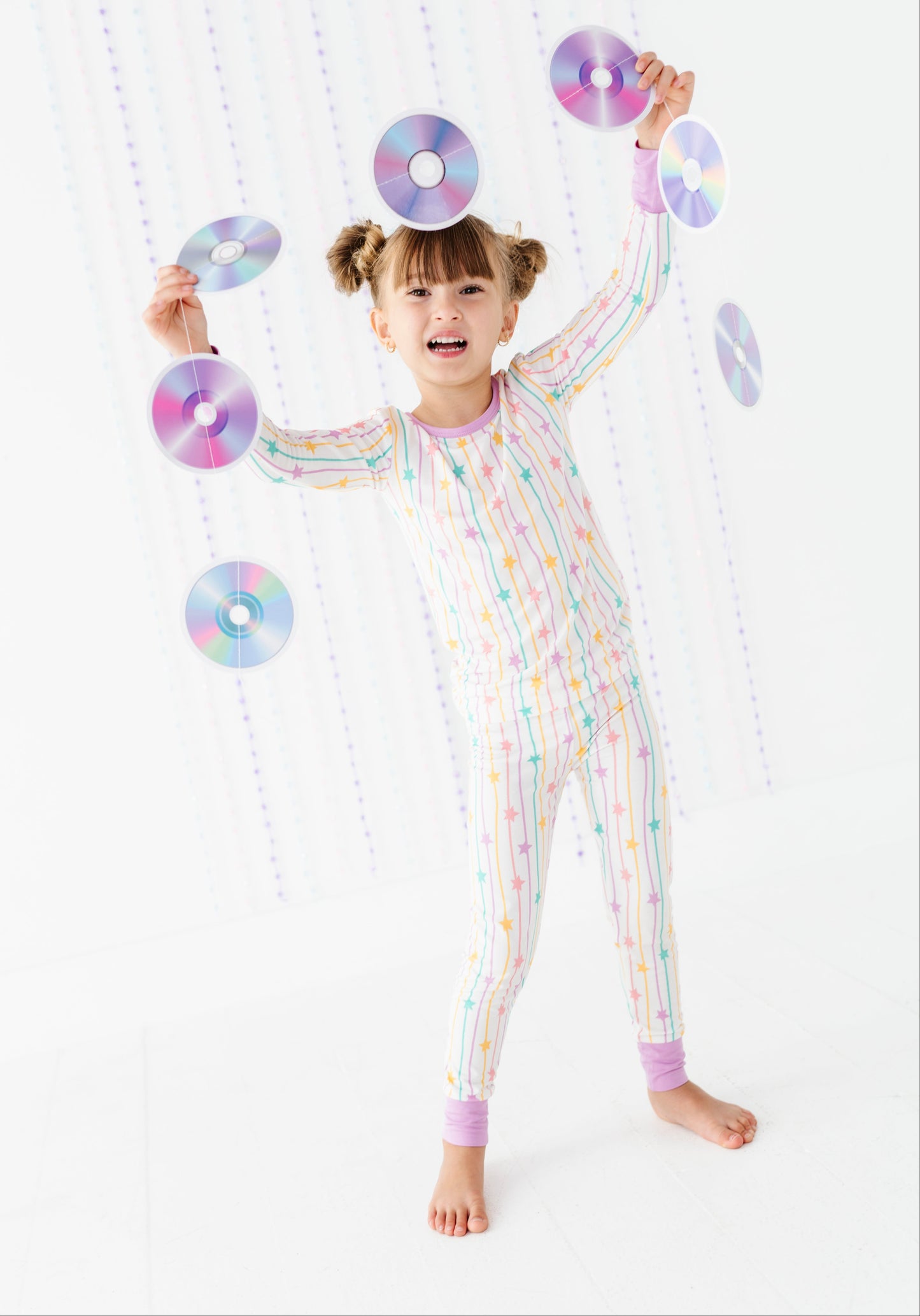 A young girl with pigtails stands barefoot in a white jumpsuit covered in pastel rainbow vertical stripes, balancing several CD-like discs around her head and arms against a white background with hanging decorative discs.