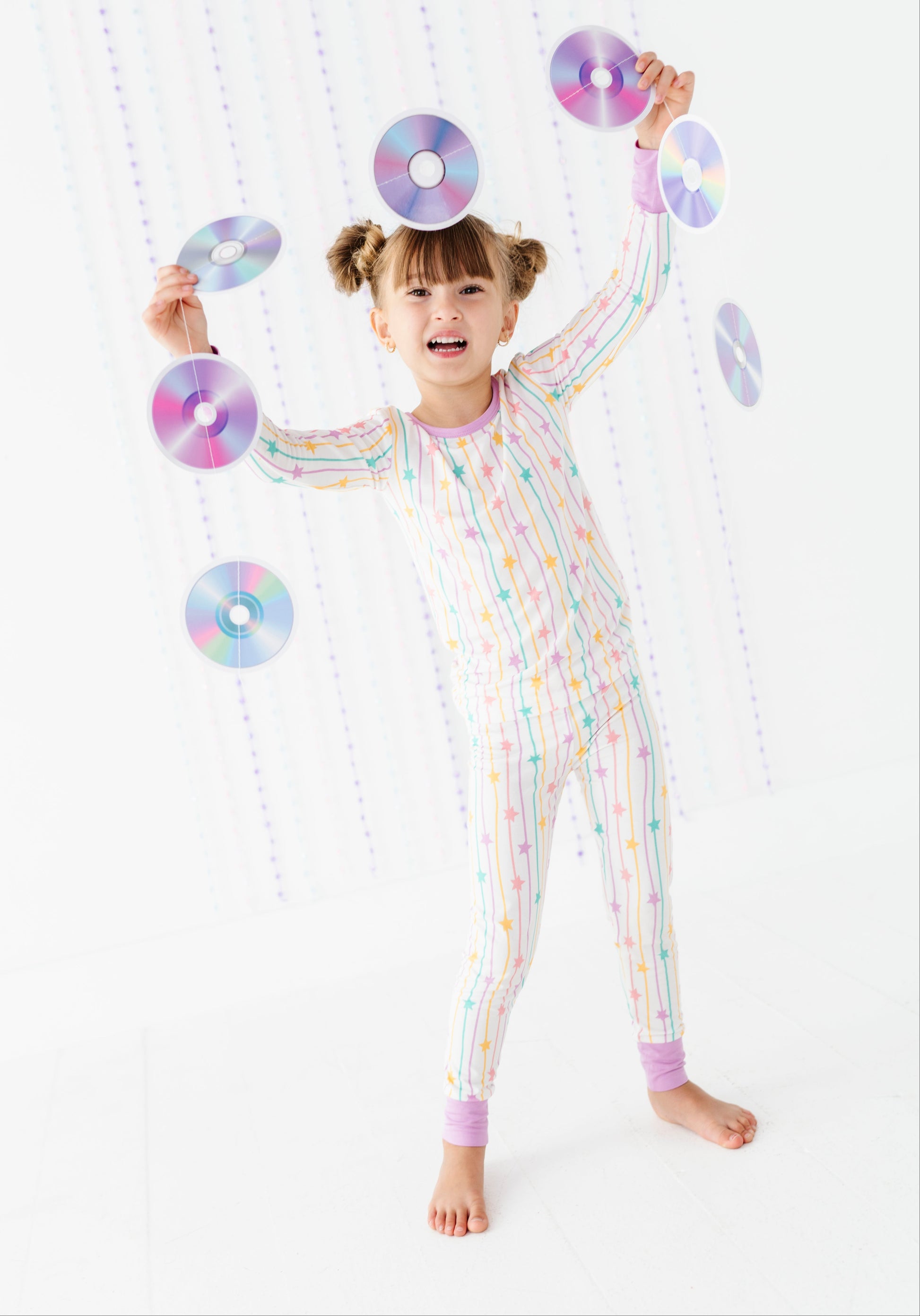 A young girl with pigtails stands barefoot in a white jumpsuit covered in pastel rainbow vertical stripes, balancing several CD-like discs around her head and arms against a white background with hanging decorative discs.