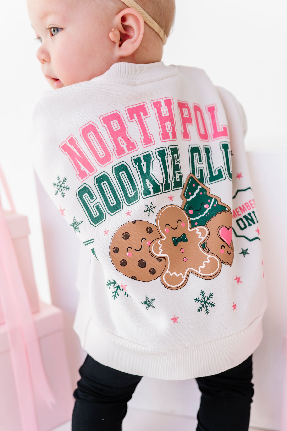 A baby wearing a white Christmas-themed crewneck sweatshirt from the back, featuring 'NORTH POLE COOKIE CLUB'.
