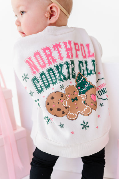 A baby wearing a white Christmas-themed crewneck sweatshirt from the back, featuring 'NORTH POLE COOKIE CLUB'.