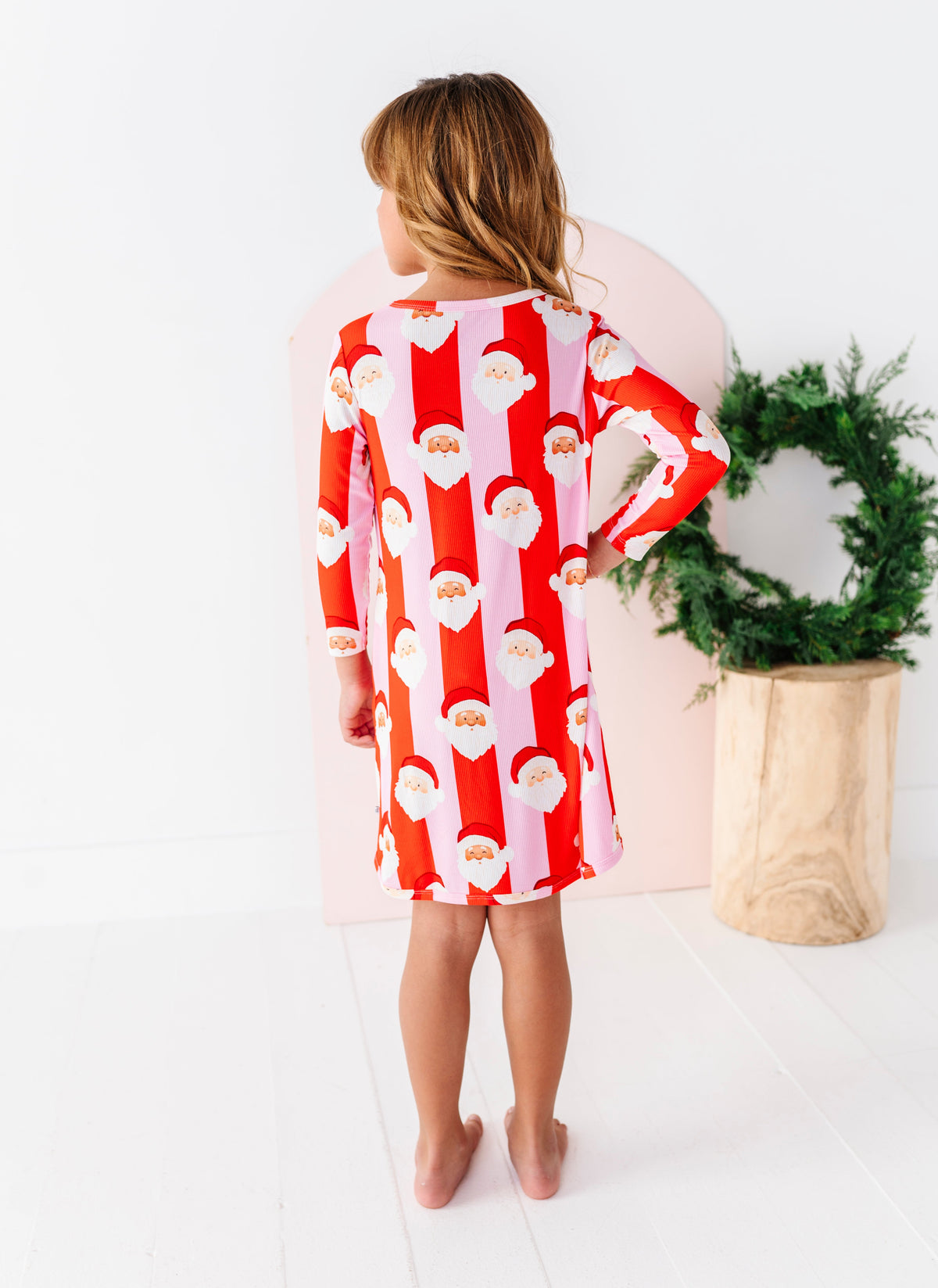 The back view of a young girl wearing a long-sleeve bamboo gown with a festive Santa Claus pattern, featuring alternating vertical stripes of red and light pink with Santa faces and beards.