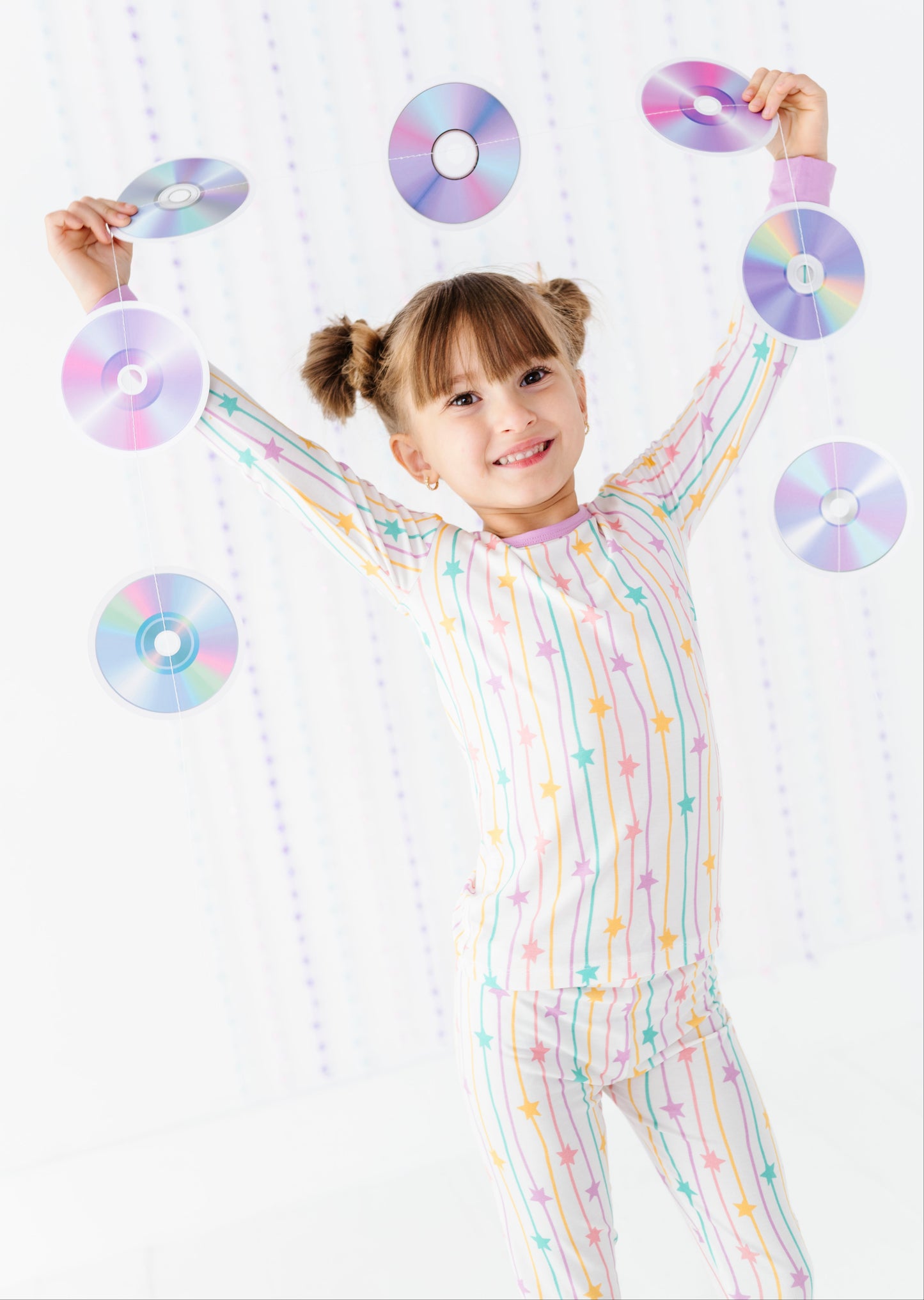 A young girl with twin buns smiles and raises her arms while wearing a pastel rainbow striped and star-patterned one-piece pajama set. She is holding several iridescent CD-ROMs against a bright white background with a subtle vertical dotted pattern.