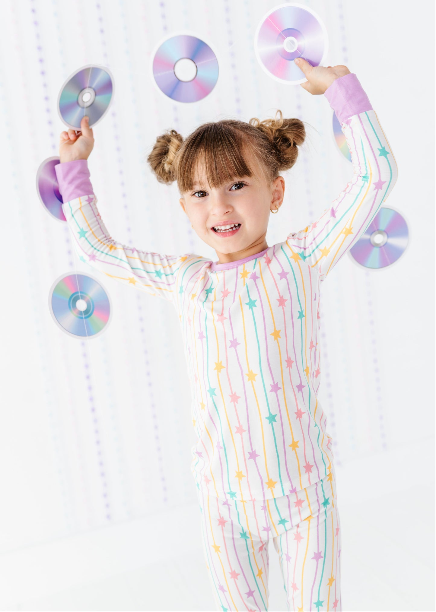 A young girl with brown pigtails smiles while balancing several reflective CD-ROMs around her head. She is wearing a pastel rainbow pajama set with vertical stripes and star patterns on a white background.