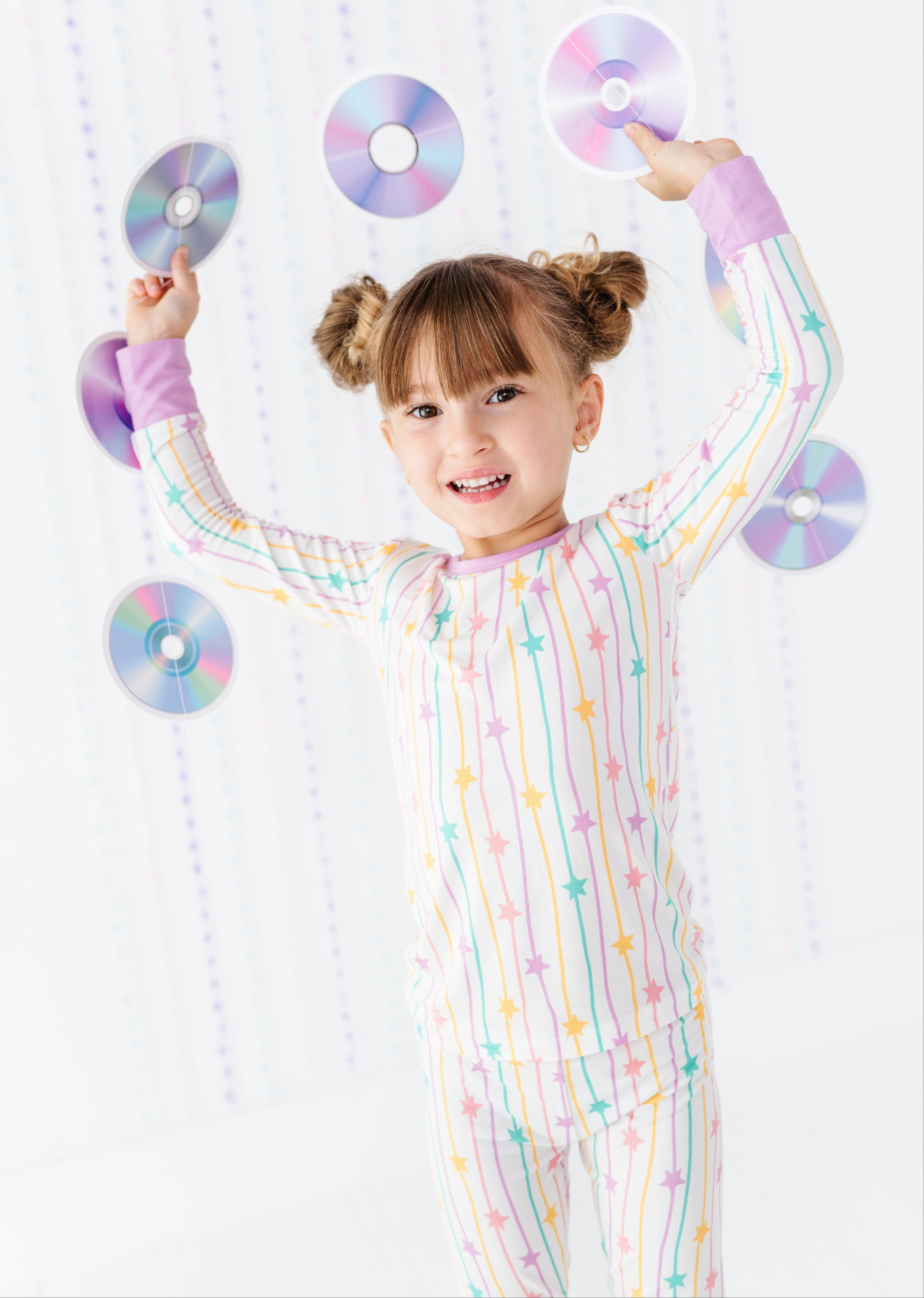 A young girl with brown pigtails smiles while balancing several reflective CD-ROMs around her head. She is wearing a pastel rainbow pajama set with vertical stripes and star patterns on a white background.