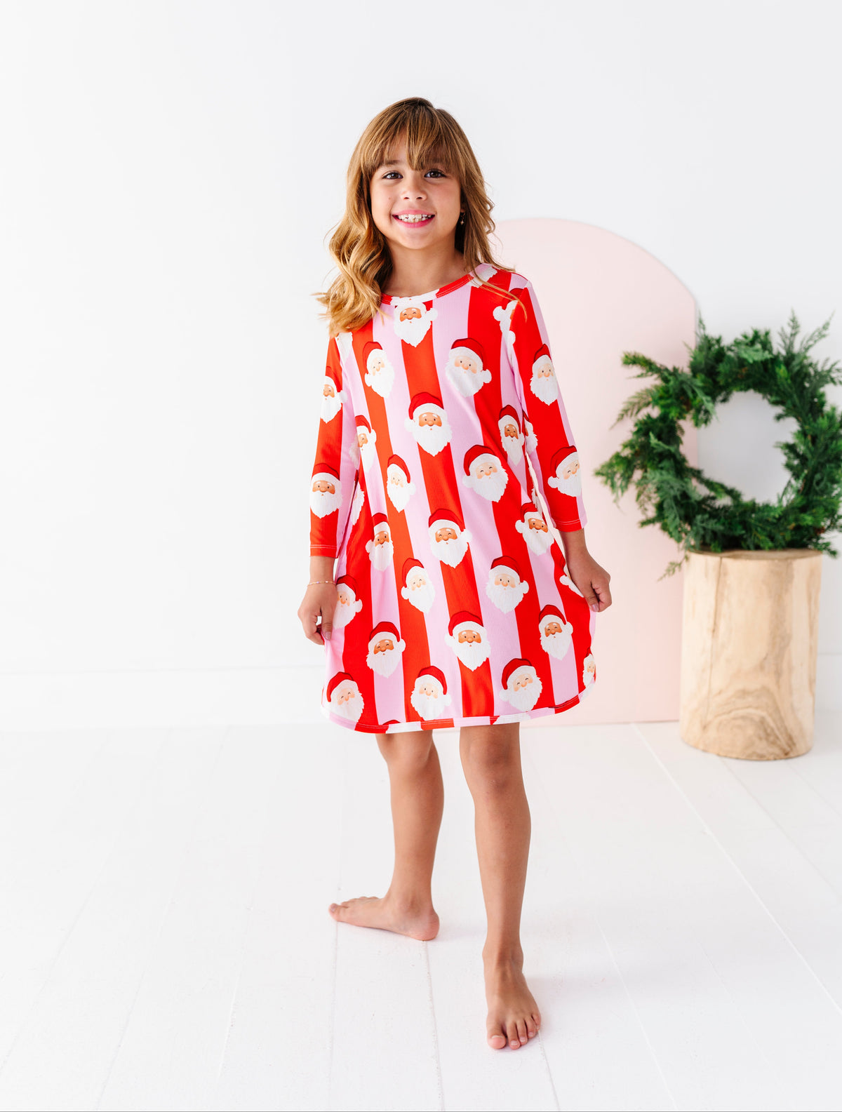 A young girl stands barefoot in a bright white studio, wearing a red and white long-sleeve bamboo gown with a repeating Santa Claus face pattern. A pink circular backdrop and a green wreath in a wooden pot are behind her.