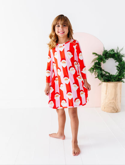 A young girl stands barefoot in a bright white studio, wearing a red and white long-sleeve bamboo gown with a repeating Santa Claus face pattern. A pink circular backdrop and a green wreath in a wooden pot are behind her.
