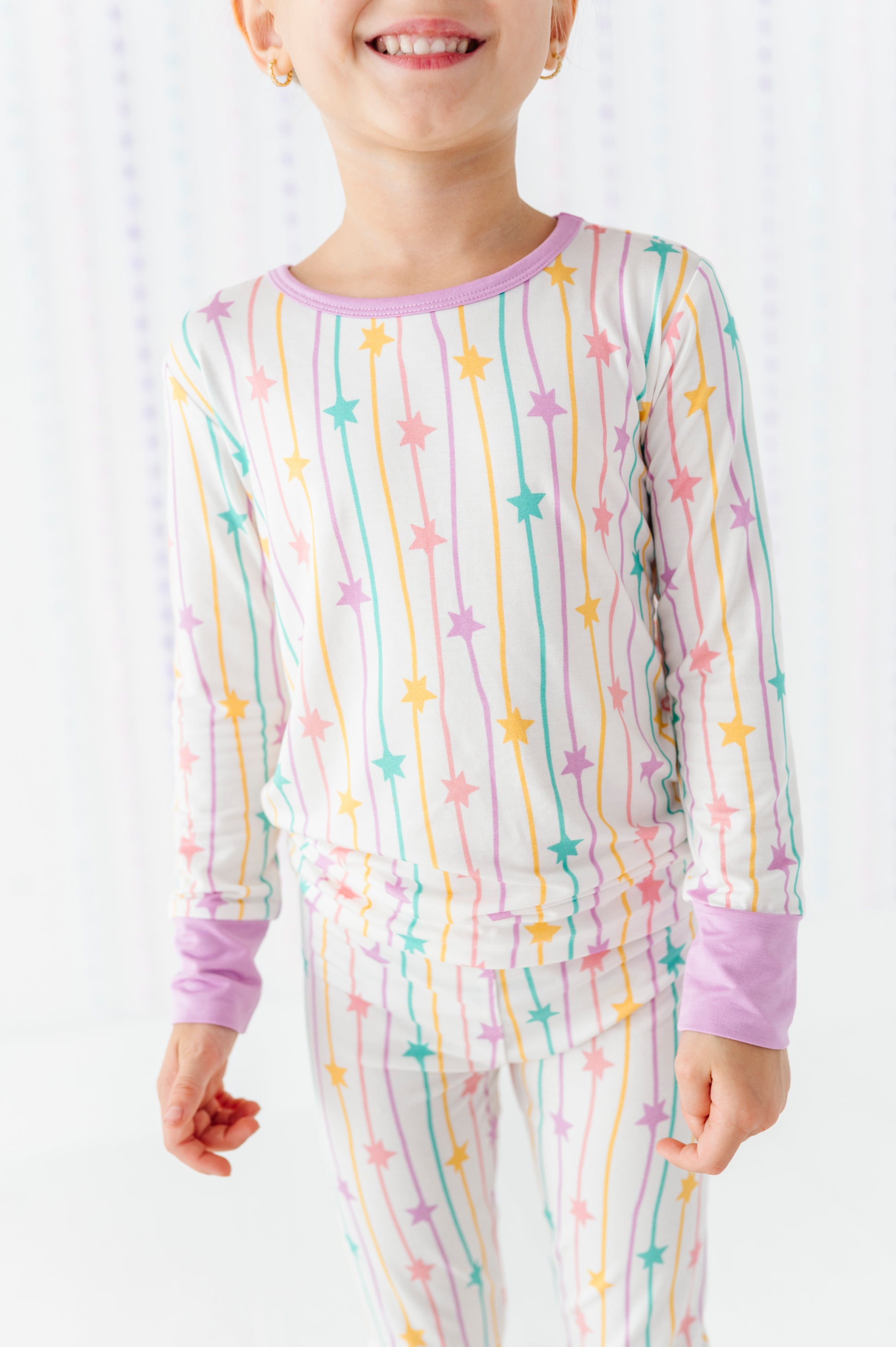A child wearing a long-sleeve pajama top and matching pants with a pastel rainbow star-and-stripe print on a white background.