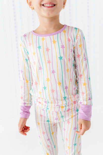 A child wearing a long-sleeve pajama top and matching pants with a pastel rainbow star-and-stripe print on a white background.