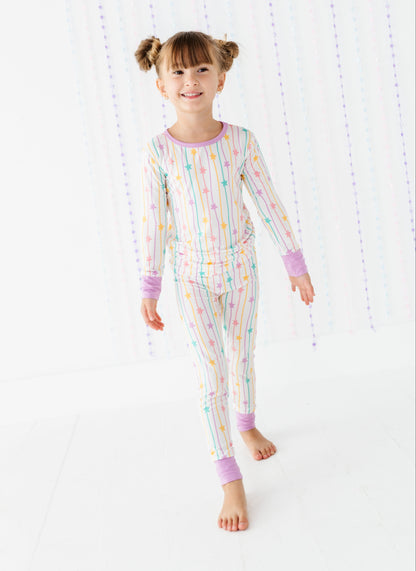 A young girl with her hair in two buns is wearing a long-sleeve pajama set with pastel rainbow stripes and small star patterns. She is standing barefoot against a bright white background with hanging decorative beads.