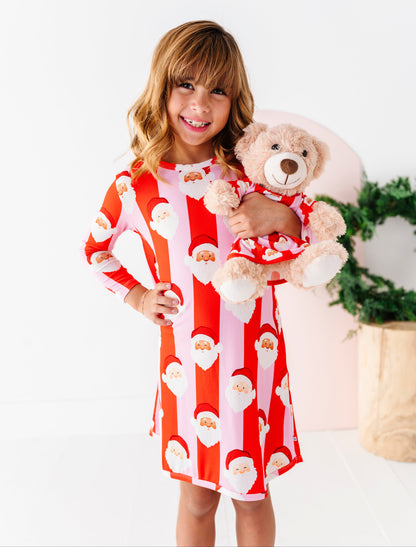 A young girl wearing a long-sleeve bamboo gown with a festive Santa Claus pattern, featuring alternating vertical stripes of red and light pink with Santa faces and beards. She's holding a teddy bear wearing a matching red and light pink gown. 